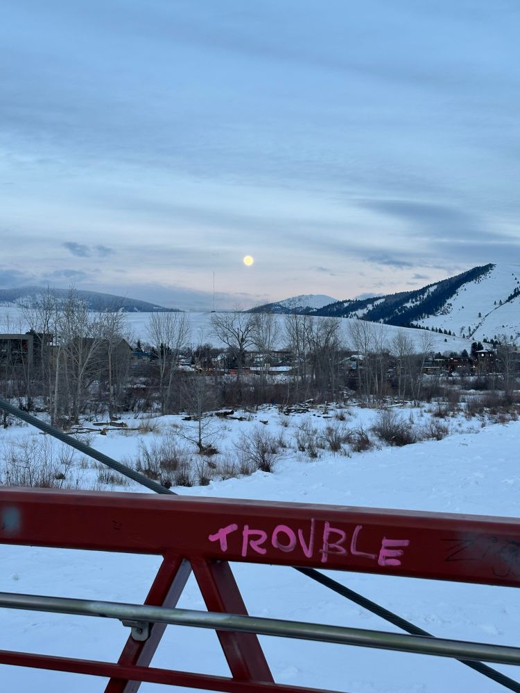 Image of a Montana winter landscape showing some whisper clouds. The moon is rising over the mountains and, in the foreground, there is the guard rail of a bridge with the word “trouble” written on it in pink paint. 