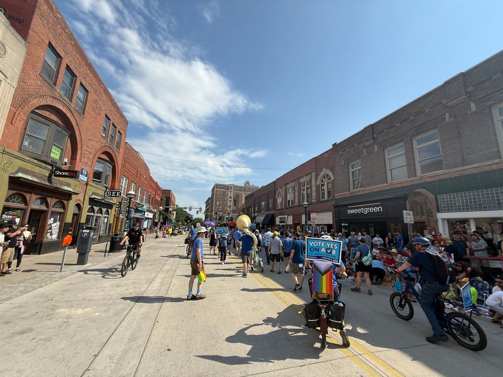 A picture of the 4th of July parade on State St. the sunny side of the street is abandoned; a thick crowd and all the participants are on the one half of the street that has shade.  