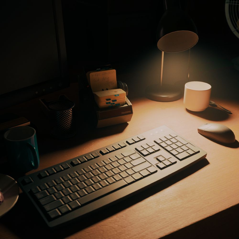 Virtual photography capture from Alan Wake 2. A desk in the office is viewed from above and at an angle, as though you were about to sit down. A single desk lamp lights the area, showing a white coffee mug, a wireless mouse, keyboard, blue coffee mug, mesh pencil cup and a rolodex