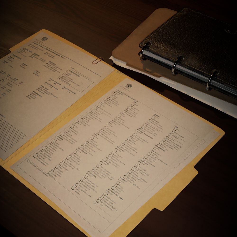 Virtual photography capture from Alan Wake 2. A "Long Term Care Assessment" is laid out in an open manilla folder on a brown wooden table. A darker folder is nearby, beneath a dark brown leather notebook that is held together with large silver rings, like you'd find in a binder.
