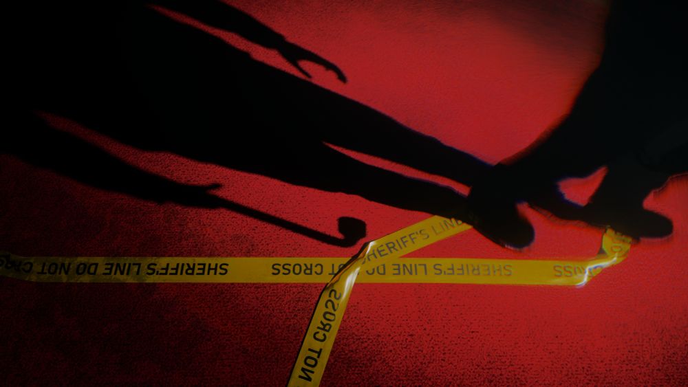 Virtual Photography capture from Alan Wake 2. A "Taken" enemy walks across a red carpet with a long shadow appearing behind them. In the shadow, it can be seen that the enemy is carrying a pipe to be used as a weapon. The enemy is walking over yellow police tape that is on the floor which reads, "Sheriff's Line Do not Cross"