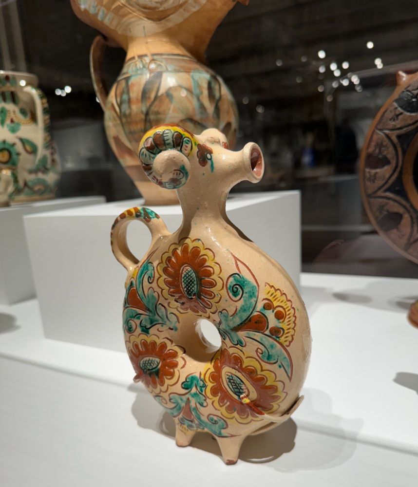 A small round teapot with a donut hole in the center. It has tiny peg feet, a thin circle tail as a handle, and a ram’s head with a pursed mouth as the pour spout. The base color is beige with big orange and yellow flowers and blue leaves painted on it. The tail and horns are similarly decorated. The overall effect is very whimsical and adorable. 