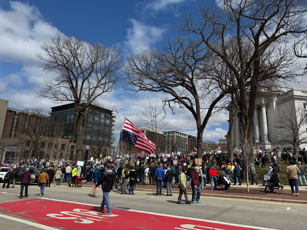 A large crowd is gathered in front of the state capitol building. Many protesters are holding signs, some have flags including the man carrying an upside down American flag walking up the street. 