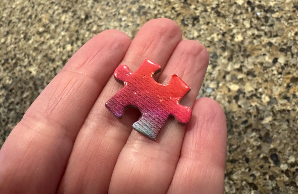 A pink puzzle piece resting in my hand, with a tiny bit of green on one foot of the piece and small dent marks across the surface. Yes, the dents are from my teeth. 