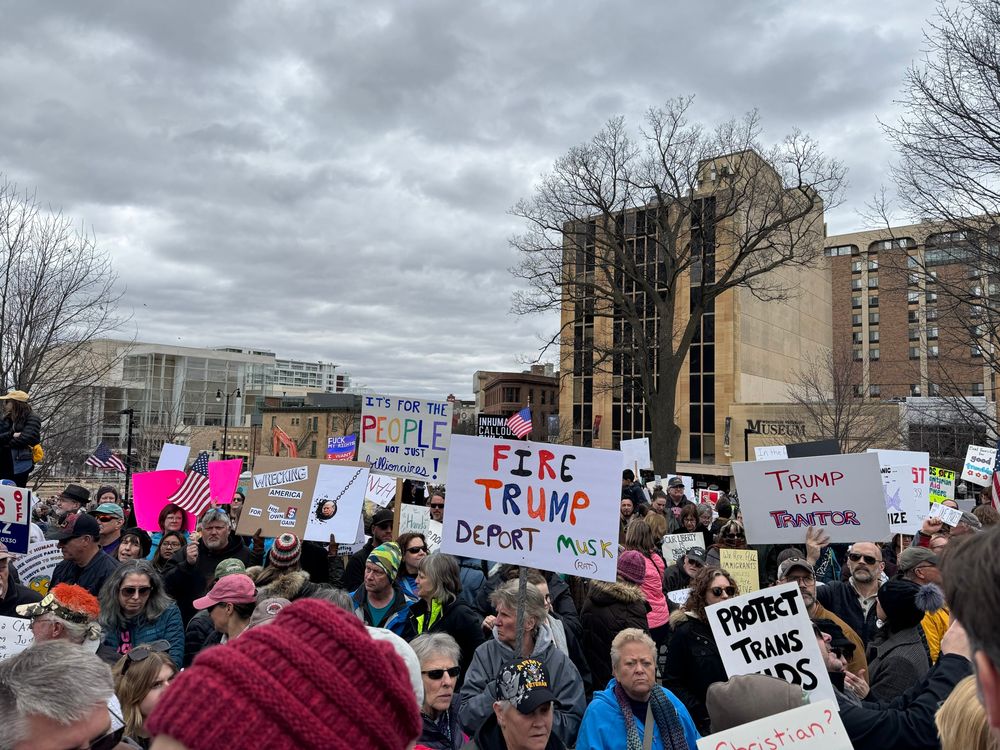 A view of the protest crowd from halfway up the stairs to the capitol building. Many signs are visible including “protect trans kids” and “Fire Trump, Deport Musk.”