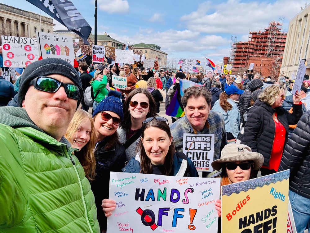 A group of friends, including me, poses in the foreground smiling and holding “hands off” posters. Many more people with flags and posters are visible in the background. This was taken at the UW campus end of State Street after we marched down the street from the state capitol building. 