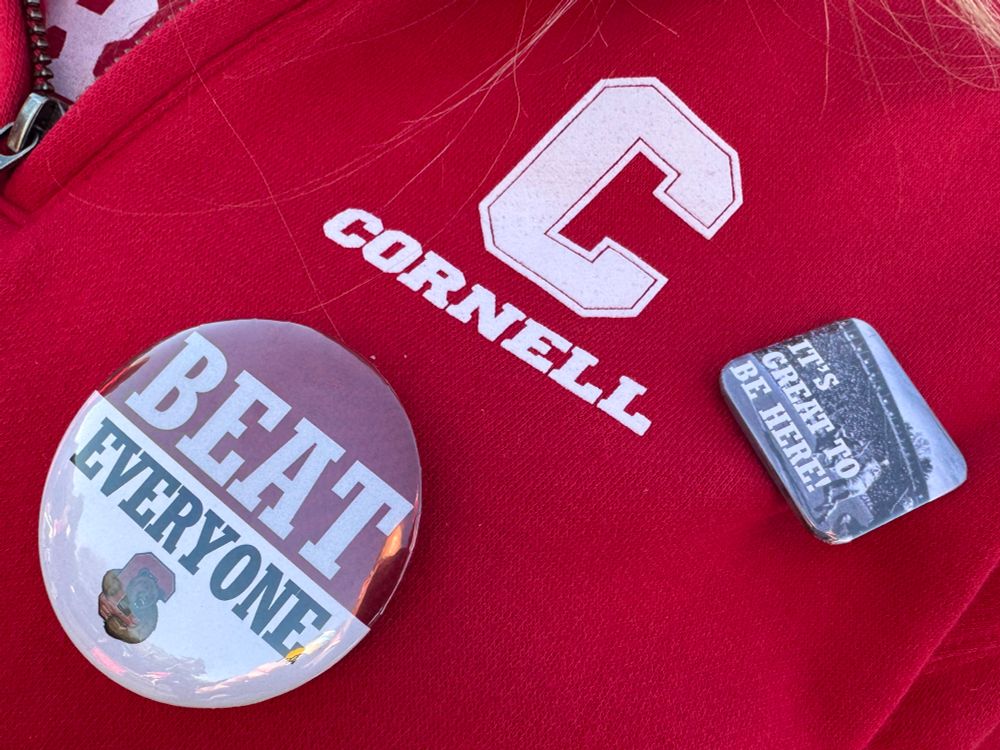 A closeup of a shirt with a C Cornell logo and two buttons. One says beat everyone with a Cornell bear logo. The other says it’s great to be here. 