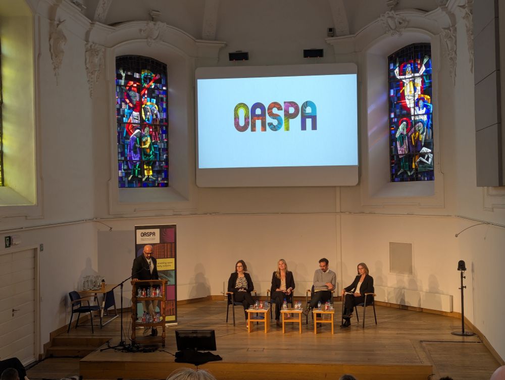 A panel of four on a stage, framed by stained glass windows and an OASPA slide