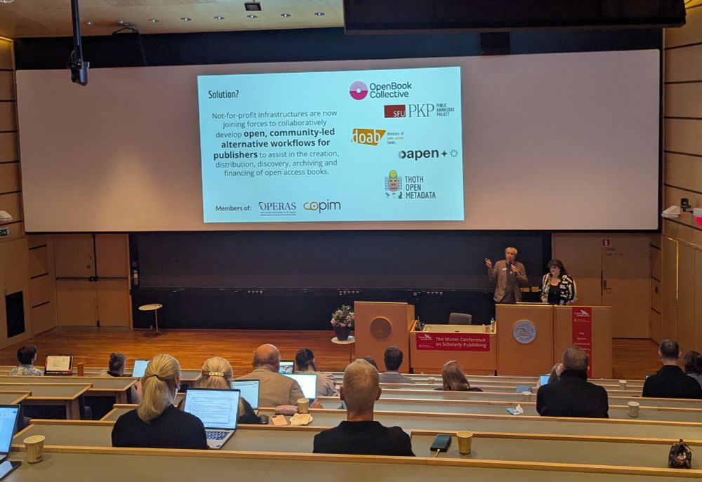 Toby gesturing to a slide above and to the left, including the logos of Open Book Collective, PKP, DOAB/OAPEN and Thoth Open Metadata. Standing next to Zoe, at the front of an auditorium