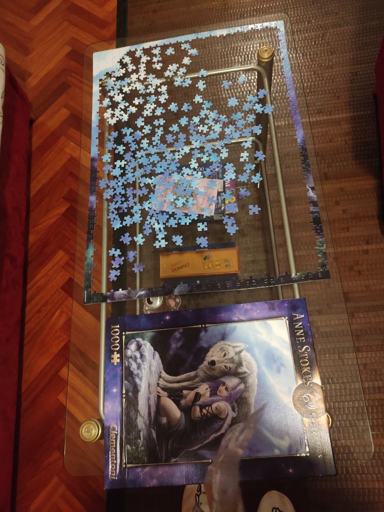 Foto de la mesa de mi salón donde se ve que hemos hecho los bordes del puzzle y separado piezas azules para hacer el cielo. Abajo se ve el dibujo del puzzle: es una ilustración de Anne Stokes, se ve a una chica peliazul con un lobo blanco