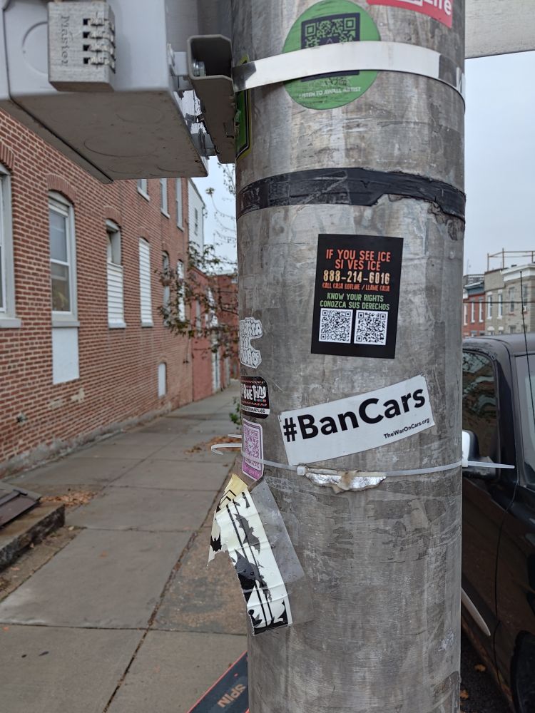 a light pole with a bunch of stickers on it, specifically two, one about how to spread the word about ICE, and the second one that reads "#BanCars"