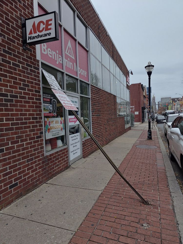 a no parking sign very bent across the sidewalk leaning into the ace hardware storefront 