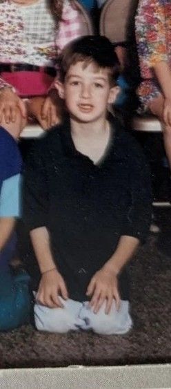 a cropped photo of an elementary school picture day showing a kid in jeans and a way too big collared shirt kneeling