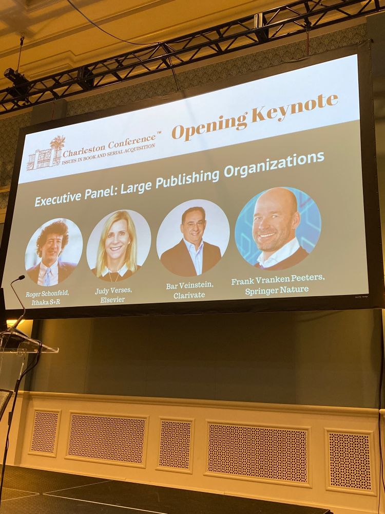 Conference screen showing a slide of the open plenary session of the Charleston Conference including the moderator: Roger Schonfeld, ITHAKA; Judy Verses, Elsevier; Bar Veinstein, Clarivate; and Frank Vranken Peeters, SpringerNature