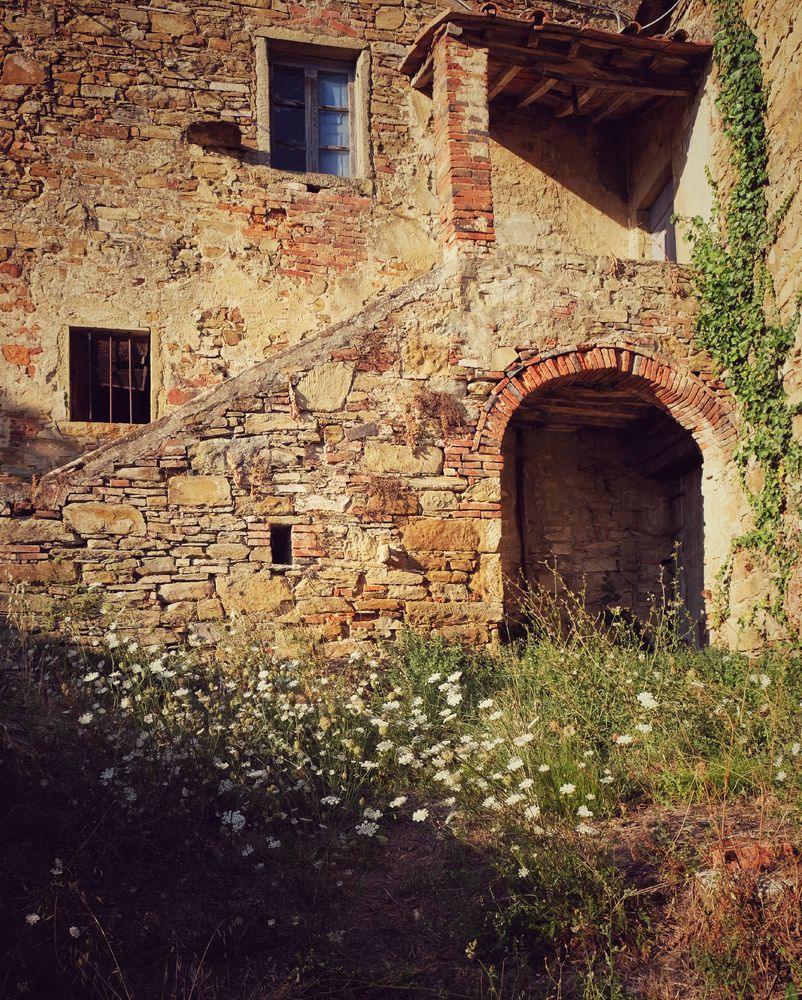 An old stone house in Toscany with an arc shaped door and small windows. Some trees are growing around the house. In front a metal table and chairs.