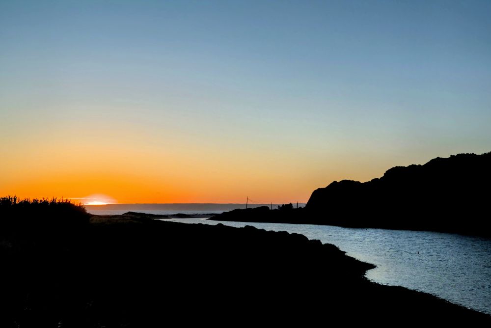 Ein Sonnenuntergang über dem Meer, umgeben von dunklen Silhouetten von Landmassen auf beiden Seiten, wobei die Sonne als leuchtender orangefarbener Streifen am Horizont erscheint.