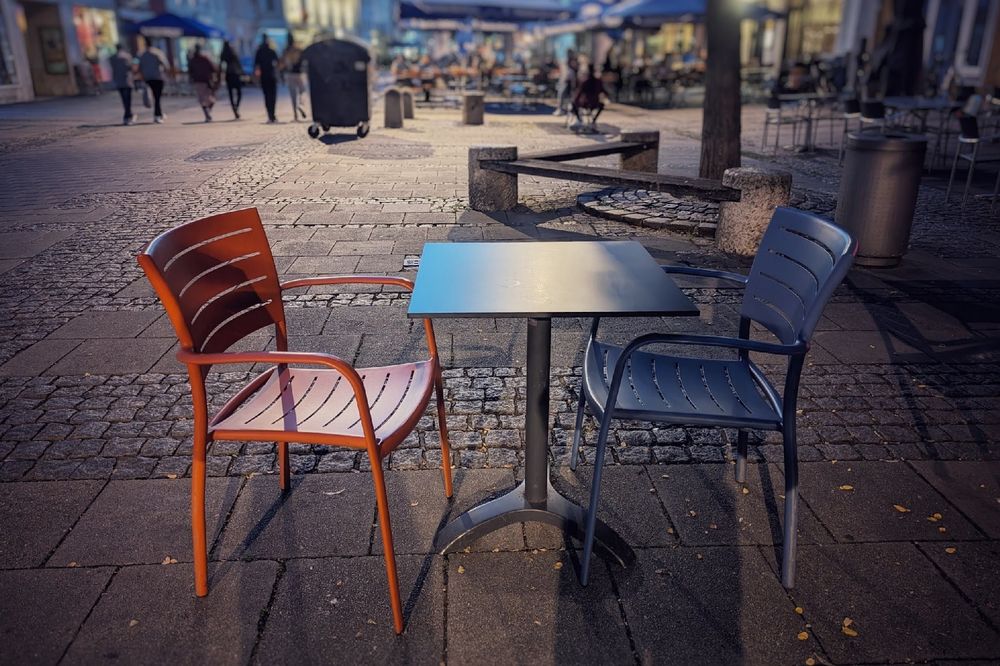 A photo in the evening shows an orange chair and a blue chair standing left and right  of a metal table. Persons are walking in the back