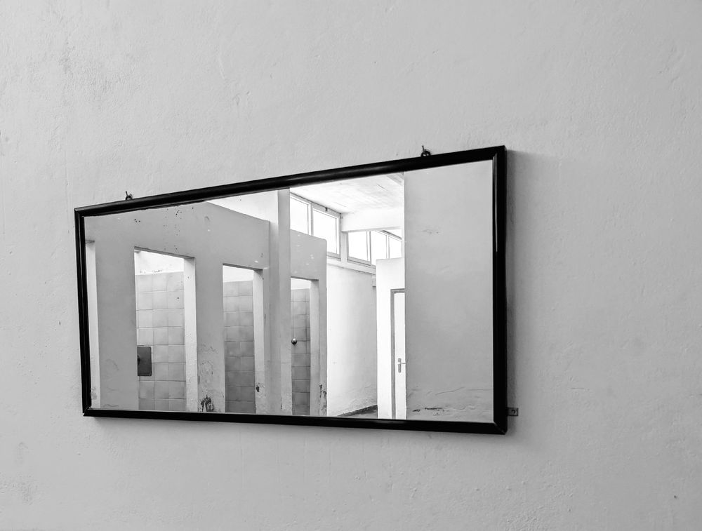 A horizontal, framed mirror hangs on a plain, light-colored wall. The mirror reflects an empty, tiled room with multiple doorways and a band of windows near the ceiling, suggesting an old, possibly abandoned changing room or bathroom. The photograph is in black and white.