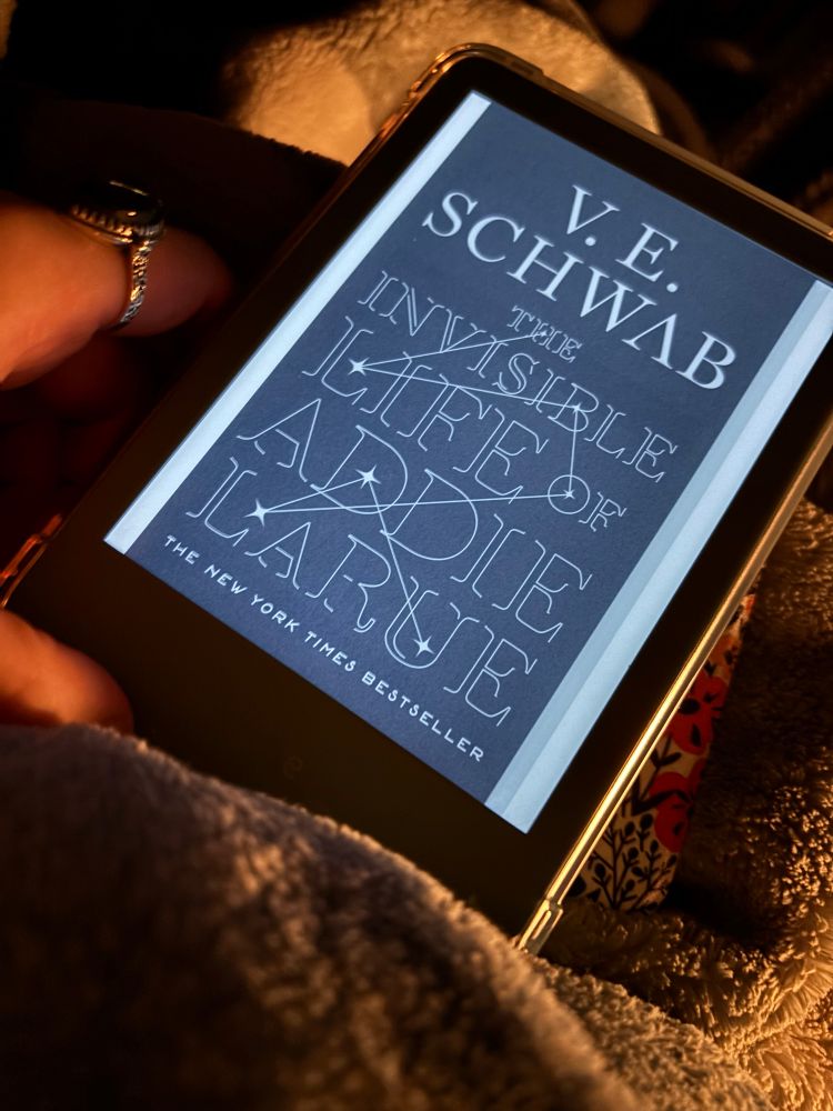 A kindle with the cover of “The Invisible Life of Addie Larue” displayed set on top of a blanket. The cover has the author’s name V.E. Schwab up top in large capital letters, and the rest of the cover is filled with the title in large capital letters. Connecting some of the letters together is a 7 point star constellation.