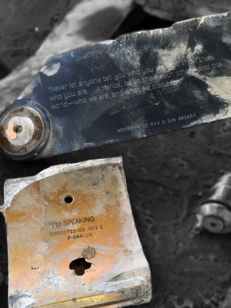 Fragments of a bomb on the ground.  The first is a long piece of metal with a quote digitally added which says "Never let anyone tell you who you are. You show them who you are. America, let us show each other--and the world--who we are, and what we stand for."  The second, a smaller piece of concrete, just has the words "I'm speaking" added in.