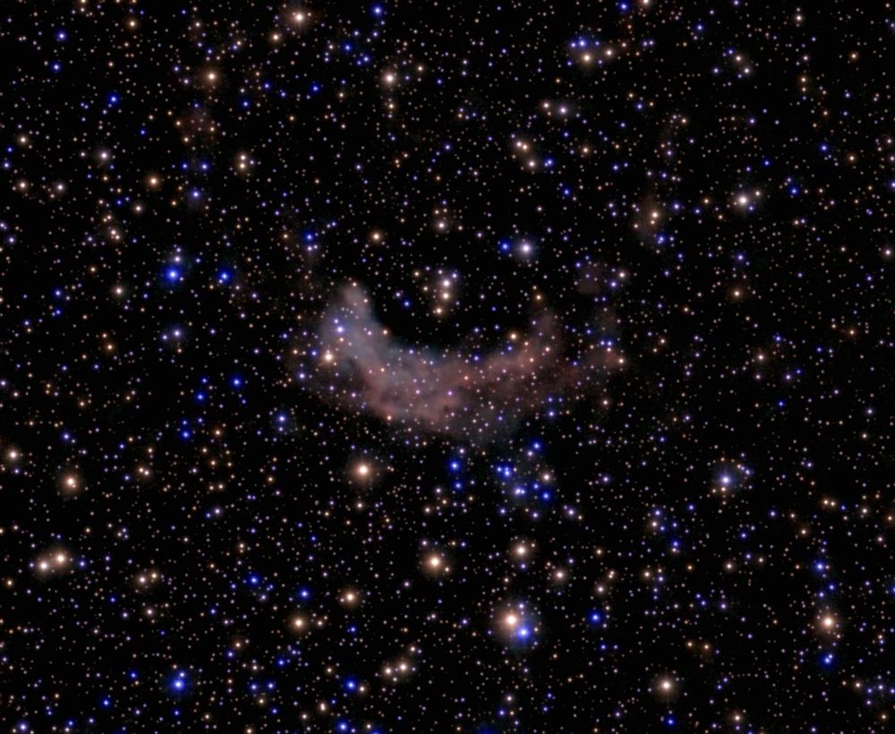 Astronomy picture of a thousand stars, mainly yellow and blue, with a banana-shaped nebula (cloud of gas and dust).