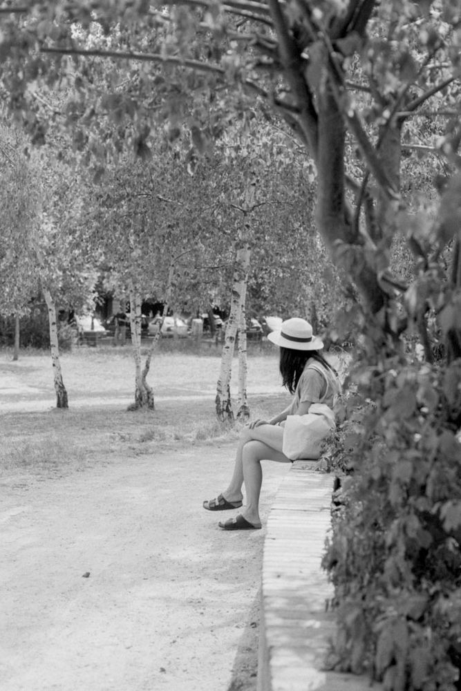 Das Schwarz-Weiß-Foto zeigt eine ruhige, sommerliche Szene in einem Park.

Eine **Frau mit hellem Sonnenhut** sitzt am rechten Bildrand auf einer niedrigen Mauer oder Bank. Sie trägt ein **helles Kleid oder eine Bluse mit Rock** und **Sandalen**, die Beine locker ausgestreckt. Ihr Blick ist nach links gewandt – ins Offene, in einen Weg, der sich zwischen **Birkenbäumen** hindurchzieht.

Die Bäume im Hintergrund und die Blätter im Vordergrund rahmen das Bild weich ein und erzeugen Tiefe. Das Licht ist mild und gestreut, vielleicht durch eine leichte Bewölkung oder das Blätterdach.

Die Atmosphäre ist **still, nachdenklich und poetisch** – fast wie eine Erinnerung. Das Bild fängt einen Moment der Pause ein, in dem Zeit und Bewegung kurz stillstehen, und verbindet Natur, Ruhe und eine zarte menschliche Präsenz zu einer harmonischen Komposition.
