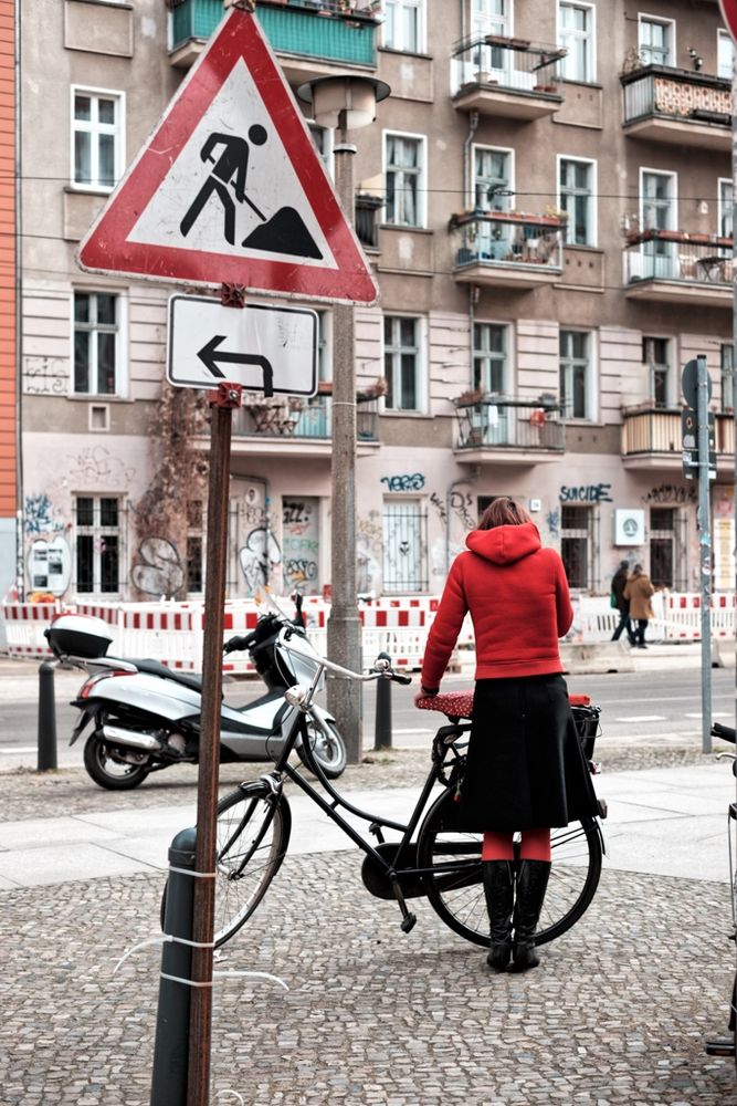 Eine Frau in roter Jacke, roter Strumpfhose und schwarzem Rock steht auf einem Bürgersteig neben ihrem Fahrrad, das an einen Pfosten gelehnt ist. Vor ihr ragt ein Baustellenschild mit dem Symbol eines Arbeiters und ein Richtungspfeil nach links in den Vordergrund. Im Hintergrund sieht man eine Berliner Häuserfassade mit Balkonen, Graffiti und einer abgesperrten Baustelle entlang der Straße. Der Farbkontrast zwischen dem kräftigen Rot ihrer Kleidung und den gedämpften Tönen der Umgebung zieht den Blick auf sie – ein Moment urbaner Stille zwischen Bewegung, Routine und Stadtchaos.