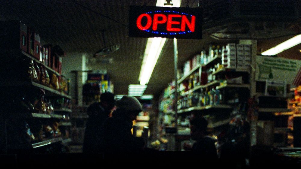 Ein dunkles, stimmungsvolles Foto zeigt das Innere eines kleinen Ladens oder Spätis. Über den Gängen hängt ein leuchtendes „OPEN“-Schild in roten und blauen Neonfarben, das den einzigen klaren Lichtakzent setzt. Darunter sind Regale mit dicht gestapelten Waren zu sehen – Chips, Konserven, Flaschen –, während mehrere Personen als dunkle Silhouetten zwischen den Gängen stehen oder sich bewegen. Das Licht der Deckenröhren verläuft in der Tiefe des Raums und schafft eine schmale, helle Perspektivlinie. Die Szene wirkt still, beinahe filmisch – ein Moment zwischen Nacht, Alltag und urbanem Leuchten.