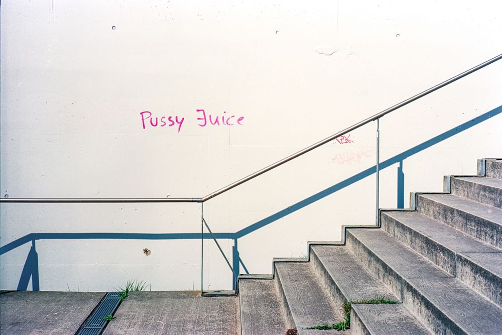 Das Bild zeigt eine minimalistische Betonszenerie mit einer Treppe auf der rechten Seite und einer weißen Wand im Hintergrund. Auf der Wand steht in pinker Schrift das Graffiti „Pussy Juice“. Ein Handlauf verläuft schräg entlang der Wand parallel zur Treppe. Der Betonboden ist sauber, aber vereinzelt wächst etwas Gras aus Ritzen. Das Licht ist hart und erzeugt klare Schatten, was auf starkes Sonnenlicht hindeutet. Das Gesamtbild wirkt still, strukturiert und urban.
