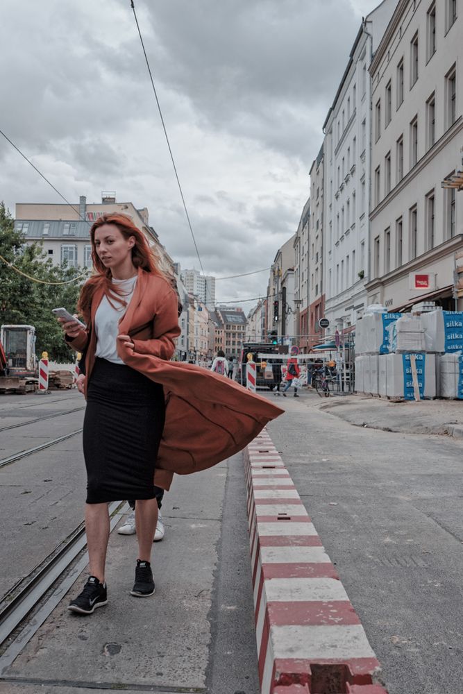 ### 🚶‍♀️ Frau mit wehendem Mantel (Farbe)

Dieses **Farbfoto** zeigt eine junge Frau mit langen, **roten Haaren**, die auf einem Bürgersteig entlang einer städtischen Straße geht. Sie trägt eine **schwarze, knielange Rock** und ein weißes T-Shirt, kombiniert mit einem **langen, orange-braunen Mantel**, dessen Saum durch eine Windböe dramatisch nach hinten weht. In der linken Hand hält sie ein Mobiltelefon.

Rechts säumt eine **niedrige Barriere** mit weiß-roten Streifen den Bürgersteig. Rechts daneben stehen **gestapelte Paletten** mit Baumaterial vor einer Reihe von **alten, hellen Mietshäusern**, die sich in die Tiefe erstrecken. Links der Frau liegen **Straßenbahnschienen** auf dem Asphalt. Der Himmel ist **bedeckt und grau**. Die Szene vermittelt den Eindruck eines geschäftigen, windigen Tages in einer Großstadt.