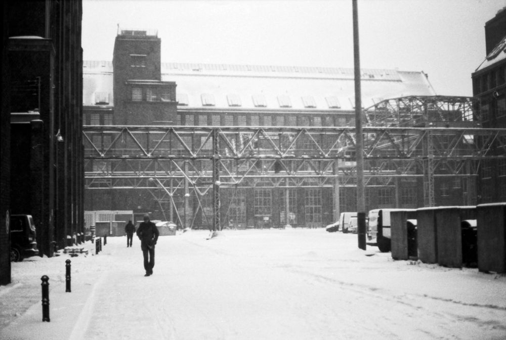 🏭 Industrielle Winterlandschaft (S/W)
Dieses Schwarz-Weiß-Foto zeigt eine breite, verschneite Straße, die durch einen Industrie- oder Werkskomplex führt. Die gesamte Szene ist von frisch gefallenem Schnee bedeckt.
Der Blick führt in die Tiefe auf ein großes, dunkles Backsteingebäude mit vielen Fensterreihen und einem turmartigen Aufbau in der Mitte. Quer vor diesem Gebäude erstreckt sich eine massive Stahlgitterkonstruktion (eine Art Förderbrücke oder Rohrleitungsgerüst), die der Szene eine starke, geometrische Textur verleiht.
Im Vordergrund führt ein Weg nach hinten. Auf diesem Weg ist eine einzelne, dunkle Person von hinten zu sehen, die in die Ferne schreitet. Links und rechts der Straße stehen dunkle Gebäude und im Vordergrund rechts einige dunkle Container oder Schuppen. Eine einzelne Straßenlaterne steht rechts im Bild. Die verschneite, körnige Szene vermittelt eine kalte, weite und ernste Stimmung.
