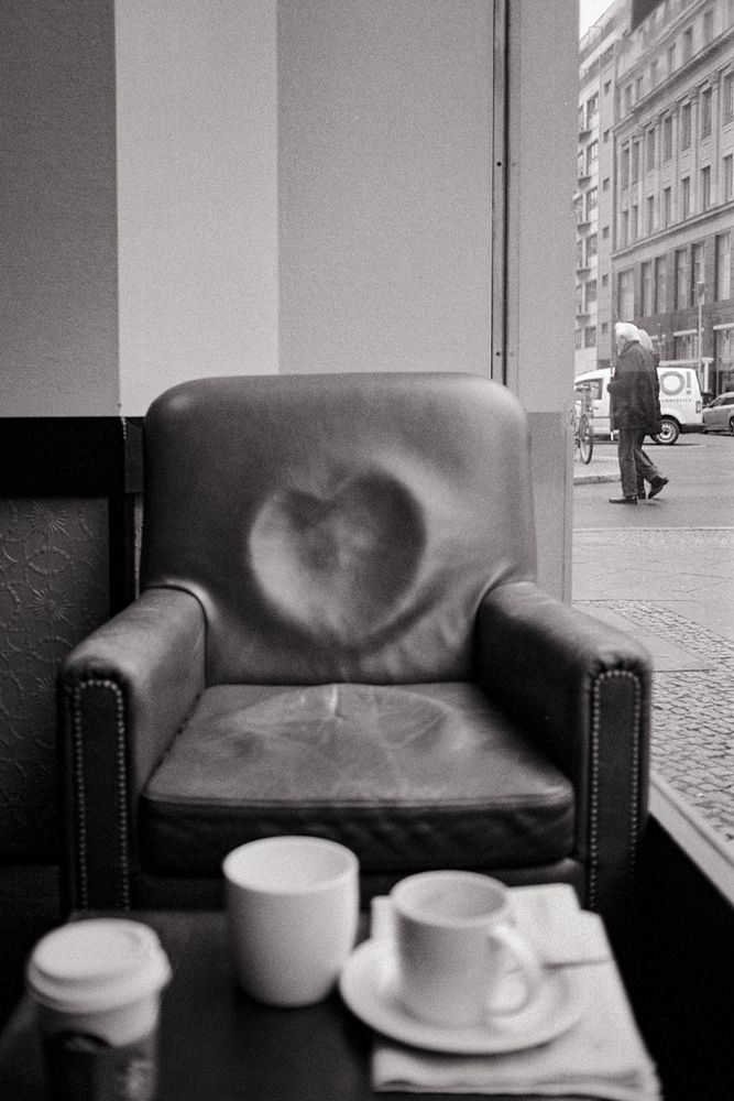 Das Schwarzweißbild zeigt einen abgenutzten Ledersessel in einem Café, auf dessen Rückenlehne sich ein herzförmiger Abrieb oder Abdruck gebildet hat – vermutlich durch häufiges Sitzen. Der Sessel steht direkt am Fenster, durch das man eine Straßenszene mit vorbeigehenden Passanten und typischer urbaner Architektur sieht. Im Vordergrund befinden sich ein Tisch mit mehreren leeren Tassen und einem Kaffeebecher, was auf kürzlich beendeten Kaffeegenuss hinweist. Die Bildkomposition wirkt ruhig und zugleich symbolisch – das „Herz“ im Sessel könnte für Wärme, Gewohnheit oder stille Begegnungen stehen. Die körnige Textur des Films verleiht der Szene eine nostalgische, intime Atmosphäre.