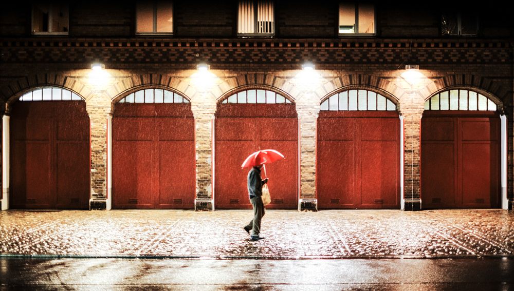 Das Foto zeigt eine nächtliche Straßenszene im Regen. Im Vordergrund geht eine Person mit einem roten Regenschirm über nasses Kopfsteinpflaster. Sie trägt eine helle Hose und eine dunkle Jacke, in der Hand hängt eine helle Tasche. Der Hintergrund besteht aus einer Fassade mit fünf großen, roten Toren, die von Backsteinbögen umrahmt werden – vermutlich eine Feuerwache. Über jedem Tor leuchtet eine helle Lampe, die den Regen sichtbar macht und die Szene stark kontrastreich ausleuchtet. Die nassen Steine am Boden reflektieren das Licht, wodurch die Szene eine atmosphärische, fast filmische Wirkung erhält.