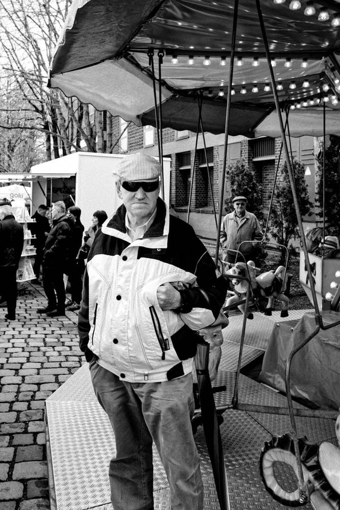 Das Schwarz-Weiß-Foto zeigt einen älteren Mann, der direkt in die Kamera blickt. Er trägt eine helle Jacke, eine karierte Schirmmütze und eine Sonnenbrille. In der linken Hand hält er einen zusammengeklappten Regenschirm, die rechte steckt lässig in der Jackentasche.

Hinter ihm steht ein kleines Karussell mit hellen Lichtern und Kinderfiguren. Im Hintergrund sieht man weitere Menschen, die sich auf dem Kopfsteinpflaster eines Marktes oder Jahrmarkts bewegen.

Die Szene wirkt spontan eingefangen, der Mann steht selbstbewusst im Vordergrund, ruhig und unbeirrt vom Trubel um ihn herum. Das Licht und die kontrastreiche Schwarz-Weiß-Darstellung verleihen dem Bild eine dokumentarische, fast zeitlose Wirkung.