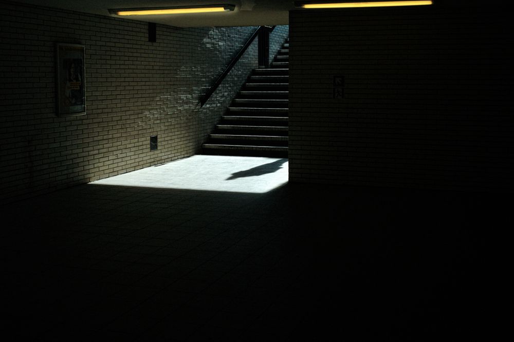 Das Bild zeigt eine fast völlig dunkle Unterführung oder U-Bahn-Passage mit Fliesenwänden und einem Treppenaufgang in der Bildmitte. Von oben fällt scharfes Sonnenlicht durch den Eingang und beleuchtet ein rechteckiges Stück Boden am unteren Ende der Treppe. In diesem hellen Ausschnitt ist der Schatten einer Person zu sehen, die gerade im Begriff ist, hinunterzugehen oder stehen bleibt – die Person selbst bleibt außerhalb des Sichtfelds. Die dominante Dunkelheit des Raums, unterbrochen vom harten Licht und dem einsamen Schatten, erzeugt eine stille, spannungsgeladene Atmosphäre mit filmischer Qualität.