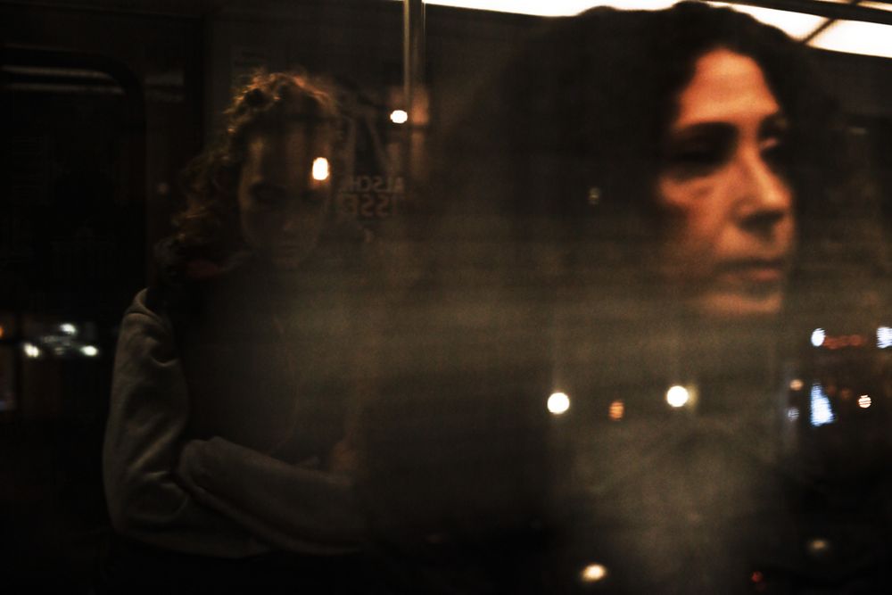 Das Foto zeigt eine dunkle, stimmungsvolle Szene in einem U-Bahn-Waggon, aufgenommen durch ein Fenster. Im rechten Bildbereich ist das unscharfe Gesicht einer Frau mit dunklen, lockigen Haaren als Reflexion im Glas zu sehen. Links steht eine weitere Person mit verschränkten Armen und gesenktem Blick, teils verdeckt durch Lichtreflexe und Spiegelungen. Mehrere helle Punkte von Innenbeleuchtung und Außenlichtern ziehen sich horizontal durchs Bild. Die unscharfen, teils überlagerten Ebenen erzeugen eine atmosphärische, leicht melancholische Stimmung, die von der Mischung aus realem Blick und Spiegelung verstärkt wird.