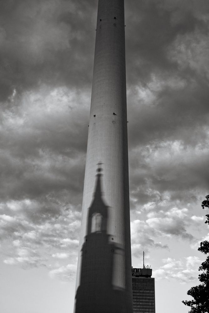 Das Bild zeigt den Berliner Fernsehturm in Schwarz-Weiß. Auf seinem schlanken Turmschaft ist der markante Schatten einer Kirche mit hohem Turm und Kreuz zu sehen, vermutlich die Marienkirche. Der Himmel ist von dramatisch wirkenden, dichten Wolken durchzogen. Rechts hinter dem Turm ist ein Hochhaus mit der Aufschrift „Park Inn“ zu erkennen. Die Szene vermittelt eine symbolträchtige Atmosphäre durch das Zusammenspiel von moderner Architektur und sakralem Schatten.

