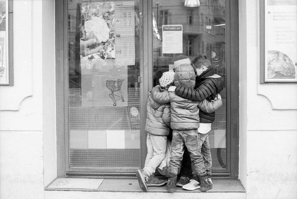 Das Schwarz-Weiß-Foto zeigt eine Gruppe von vier Kindern, die eng beieinanderstehen und sich gegenseitig umarmen. Sie tragen dicke Jacken und Mützen, was auf kühles Wetter hinweist.

Die Kinder stehen direkt vor einer Glastür eines Ladens oder Supermarktes. An der Scheibe sind Plakate und Aushänge zu sehen, unter anderem ein Bild von einem Einkaufswagen und ein Schild mit der Aufschrift „Wir stellen ein!“.

Die Szene wirkt spontan und lebendig, fast so, als hätten die Kinder ein geheimes Spiel oder einen Moment des Zusammenhalts mitten im öffentlichen Raum. Im Hintergrund spiegeln sich in der Glasscheibe Gebäude der Straße.

Das Bild vermittelt Gemeinschaft und Freundschaft, eingefangen in einer alltäglichen Umgebung.