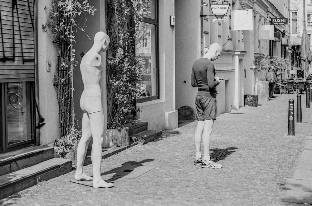 Das Schwarz-Weiß-Foto zeigt eine urbane Szene mit einem interessanten visuellen Kontrast: Links steht eine nackte Schaufensterpuppe ohne Arme vor einem Hauseingang, rechts daneben ein echter Mann in Sportkleidung, der auf sein Handy schaut. Beide stehen in ähnlicher Haltung, was auf humorvolle Weise die Grenze zwischen Mensch und Objekt verschwimmen lässt. Die gepflasterte Straße, die Schilder über den Geschäften und das helle Sonnenlicht verstärken den dokumentarischen, fast surrealen Charakter der Aufnahme. Die Szene wirkt alltäglich, aber zugleich inszeniert – ein Spiel mit Wahrnehmung und Identität.

