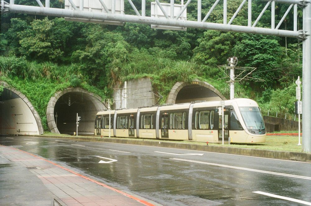 The golden TRSC tram vehicle of the Ankeng Light Rail drives out of its towards the depot. It has emerged from a tunnel cutting though a very hilly section of jungle.
