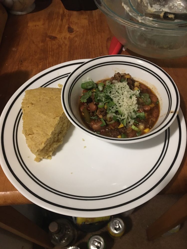 Bowl of chili topped as described on a plate next to a slice of cornbread 