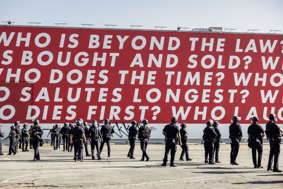 About 3 dozen LAPD officers in full riot gear, standing around looking confused, bored, aimless. A waste of taxpayer money, a threatening show of force without morality or purpose. Behind them, the north wall of the Geffen Contemporary at MOCA, covered with Barbara Kruger's "Untitled (Questions)" mural: against a field of stark red, white letters that read, in part, WHO IS BEYOND THE LAW? WHO IS BOUGHT AND SOLD? WHO DOES THE TIME? WHO SALUTES LONGEST? WHO DIES FIRST? WHO LAUGHS LAST