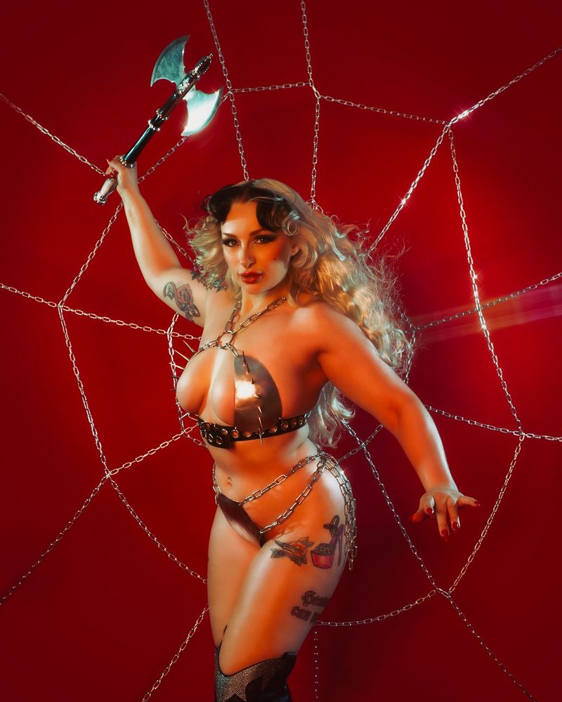 Heavy metal looking woman with long blonde curls, wielding a battle axe wearing a spiked metal bikini against a spiderweb made of chains. 