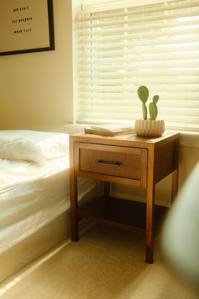 A photo of a wooden bedside table with a singular drawer. The bedside table is the focus of the piece, but beside it is a soft, white bed, and above it a window provides a stream of warm, soft light.