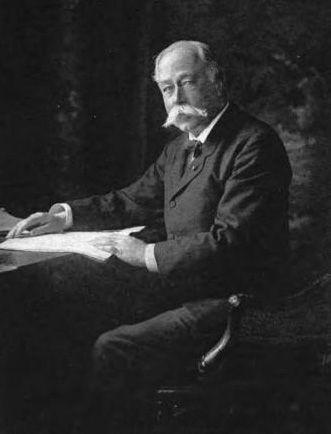Historic photograph of a man sitting at desk wearing a suit with a newspaper on the desktop.