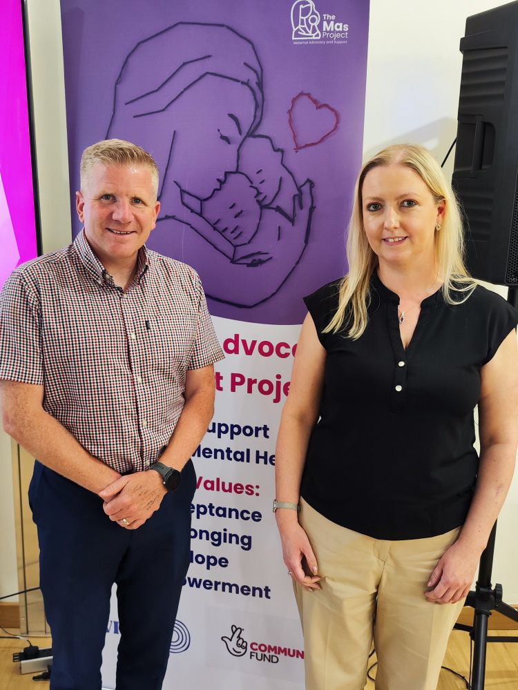 This is a photo of Commissioner Chris Quinn standing beside Clare Anderson, Maternal Advocacy & Support Project Coordinator for the Women's Resource & Development Agency. There is a purple banner behind them with the words The Mas Project on the top right hand corner.