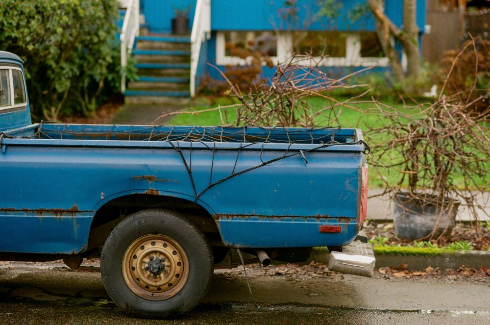 a small blue pickup, aged and gently rusting, parked in front of a small blue home on a wet day where the grass is vibrant and the concrete is shiny.