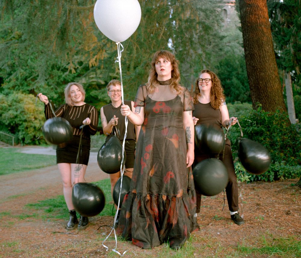 Vivian stands front and center forlornly with a single white balloon as her bandmates eagerly swing dead-hanging black balloons behind her.