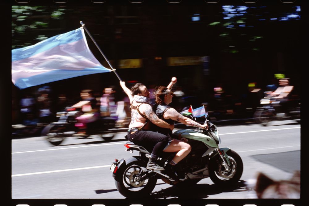two people on a sportbike speed past. the rider raises her fist, the passenger carries a trans flag high above him. the motorcycle's handlebars have Palestine and trans flags attached. 