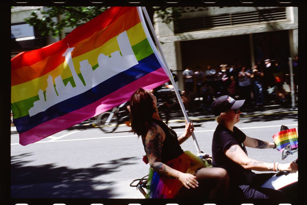 a woman on the back of a motorcycle looks up at her flag, a rainbow with a white silhouette of the Seattle skyline.